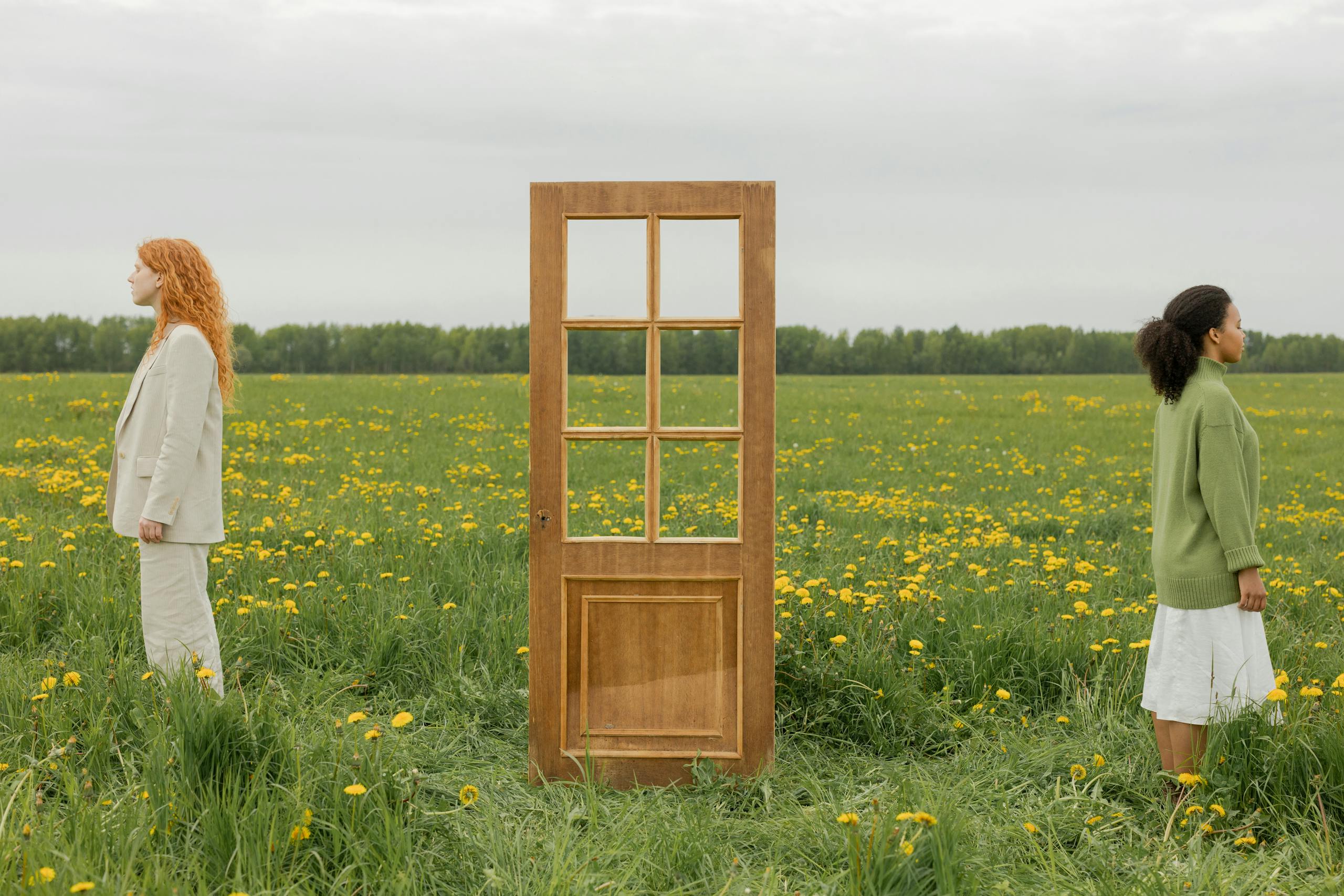 Two women standing back to back separated by a door in a field of flowers.